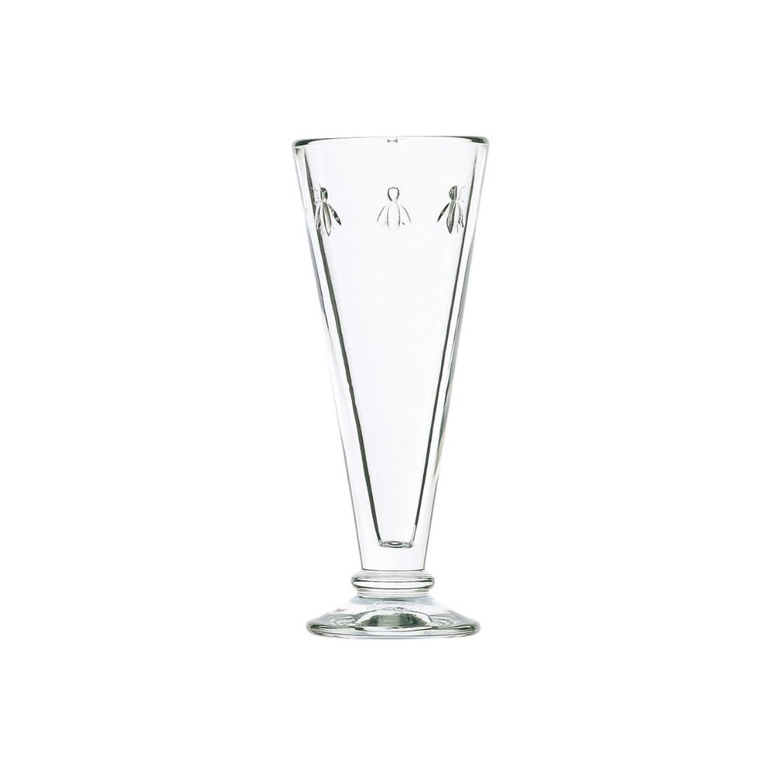 The BEE FLUTE by LA ROCHERE is a tall, clear glass vase with a narrow base and wider top, featuring an etched bee design near the rim. Its minimalist appearance with a slight tapering shape exudes French country charm, creating an elegant look reminiscent of delicate champagne flutes.