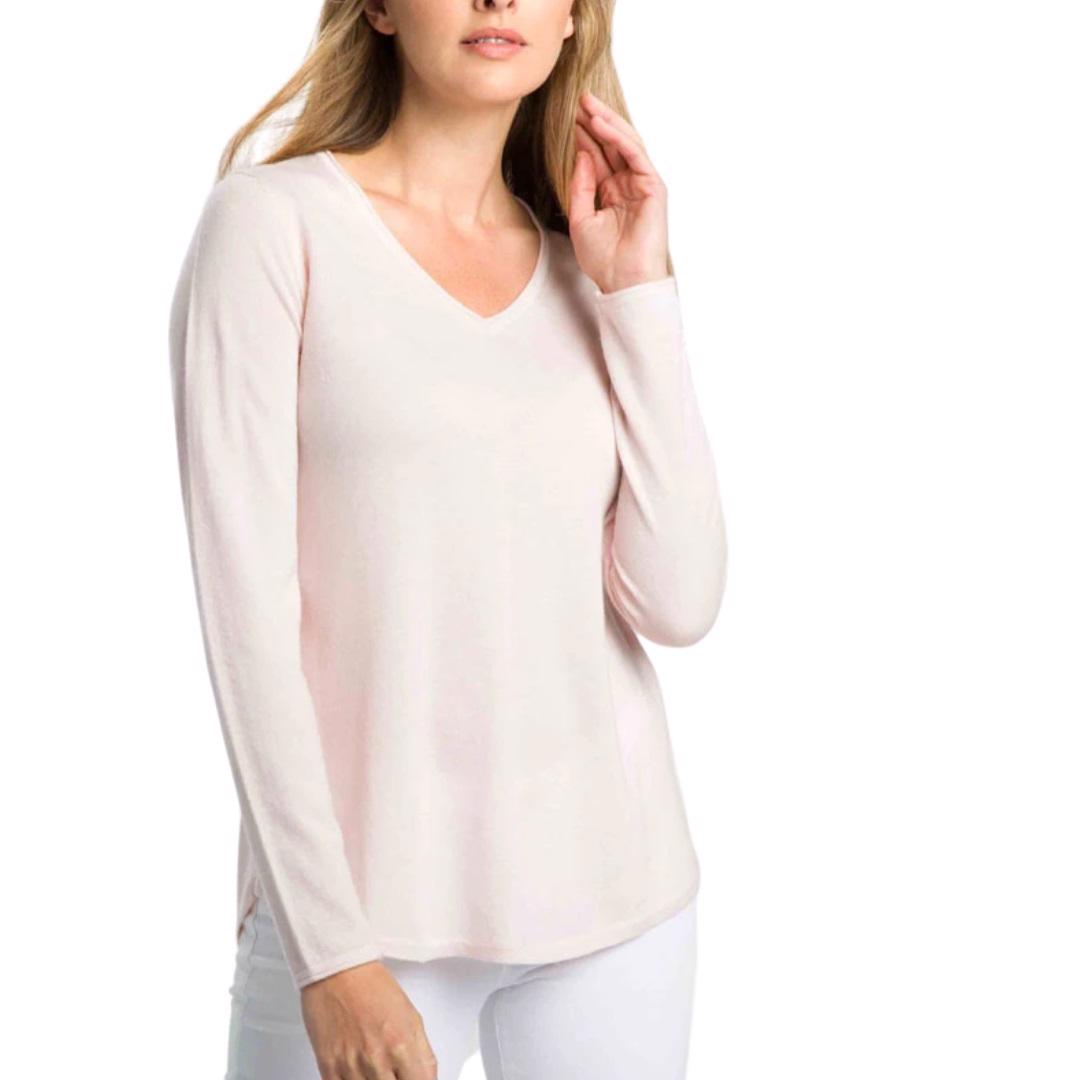 A person wearing a shell pink Kinross Cashmere - Worsted Round Hem V-neck top and white pants is touching their hair with one hand. The background is plain white.