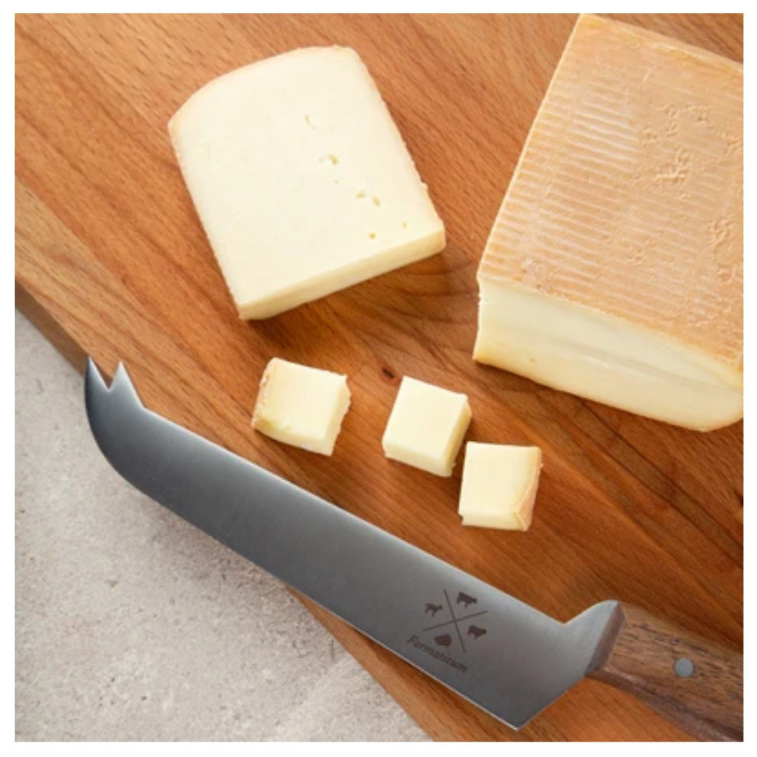 A wooden cutting board holds a block and slices of cheese, along with four cheese cubes. Nearby, a large FORMATICUM PRO TASTING CHEESE KNIFE featuring a distinctive stainless steel blade is positioned partially off the edge of the board. The scene is set on a light-colored surface.