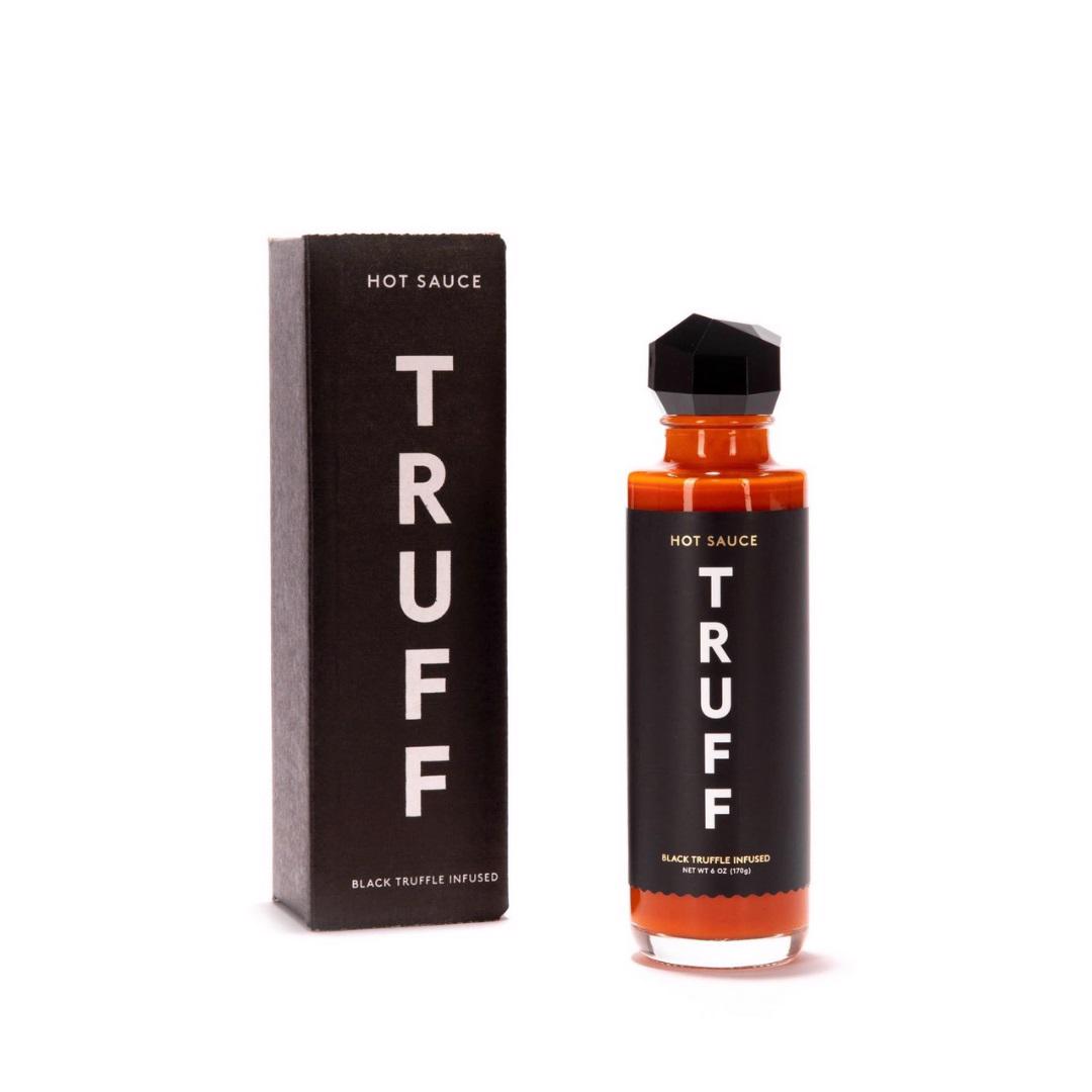 A bottle of TRUFF - HOT SAUCE with a black label and geometric black cap sits next to its matching black box packaging. The label and box text reads, "TRUFF HOT SAUCE - Black Truffle Infused." The bottle contains a vibrant red-orange sauce infused with organic agave nectar.