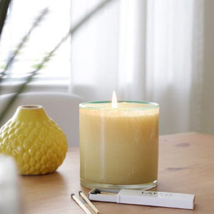 A 6.5 oz LAFCO - French Lilac, Poolhouse candle is displayed in a cylindrical glass container filled with a beige substance. The container features a subtle greenish tint at the rim, evocative of French lilac, and casts a shadow to the right, suggesting the light source is on the left. The background is plain white.