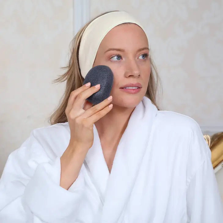A black Daily Concepts Charcoal Konjac Sponge is placed beside its packaging, labeled &quot;Daily Concepts Daily Konjac Sponge.&quot; The packaging highlights that it is made from 100% natural and preservative-free konjac root and features the word &quot;charcoal.&quot; It is organic, vegan, biodegradable, and highly effective for removing skin impurities.
