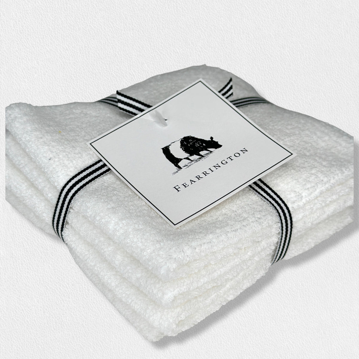 A neatly folded white textured towel, part of the FEARRINGTON LIFESTYLE TOWEL COLLECTION - WAFFLE FACE CLOTH S/4 by HARBOR LINEN, is tied with a black and white striped ribbon. Attached to the ribbon is a square tag featuring a black silhouette of a cow with &quot;FEARRINGTON&quot; in capital letters beneath it. The towel sits on a white surface.