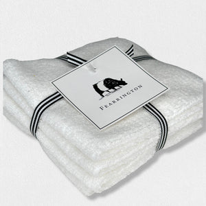 A neatly folded white textured towel, part of the FEARRINGTON LIFESTYLE TOWEL COLLECTION - WAFFLE FACE CLOTH S/4 by HARBOR LINEN, is tied with a black and white striped ribbon. Attached to the ribbon is a square tag featuring a black silhouette of a cow with "FEARRINGTON" in capital letters beneath it. The towel sits on a white surface.