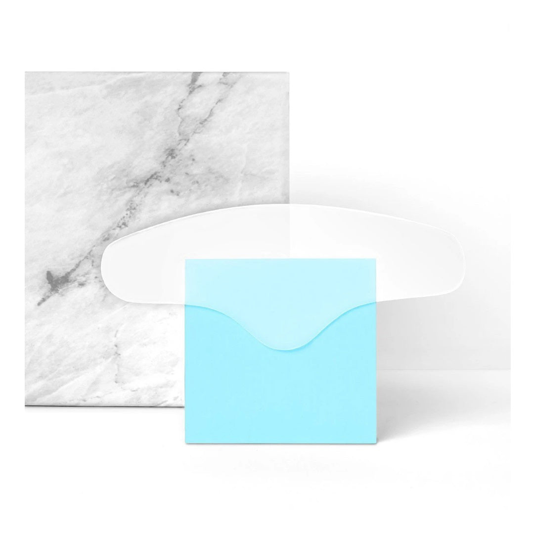 A minimalist arrangement featuring a white marble-patterned notebook, SIO BEAUTY&#39;s transparent curved SIO BROWLIFT resembling medical grade silicone patches, and a small blue envelope. The marble notebook has subtle gray veining, and the blue envelope is centered in front of the notebook.