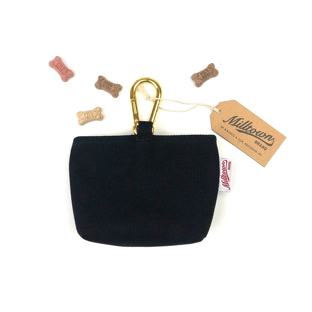 The DOG TREAT CASE (ONYX) from MILLTOWN BRAND boasts a minimalist black fabric design, featuring a golden carabiner clip at the top and the iconic Milltown Brand label. Small, bone-shaped dog treats in various colors are scattered around the pouch.