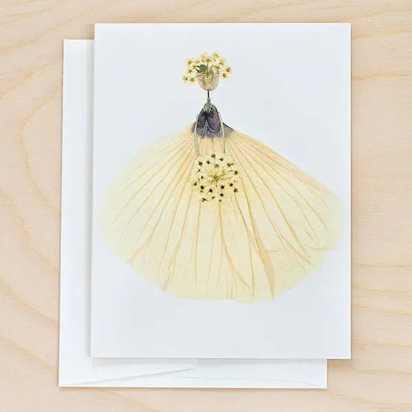 The PETAL PEOPLE PRESS - BRIDE CARD by PETAL PEOPLE PRESS, displayed on a wooden surface, showcases an elegant dress crafted from pressed white flowers and petals. The nature-inspired design resembles the silhouette of a bride in a gown, holding a bouquet of small white flowers.