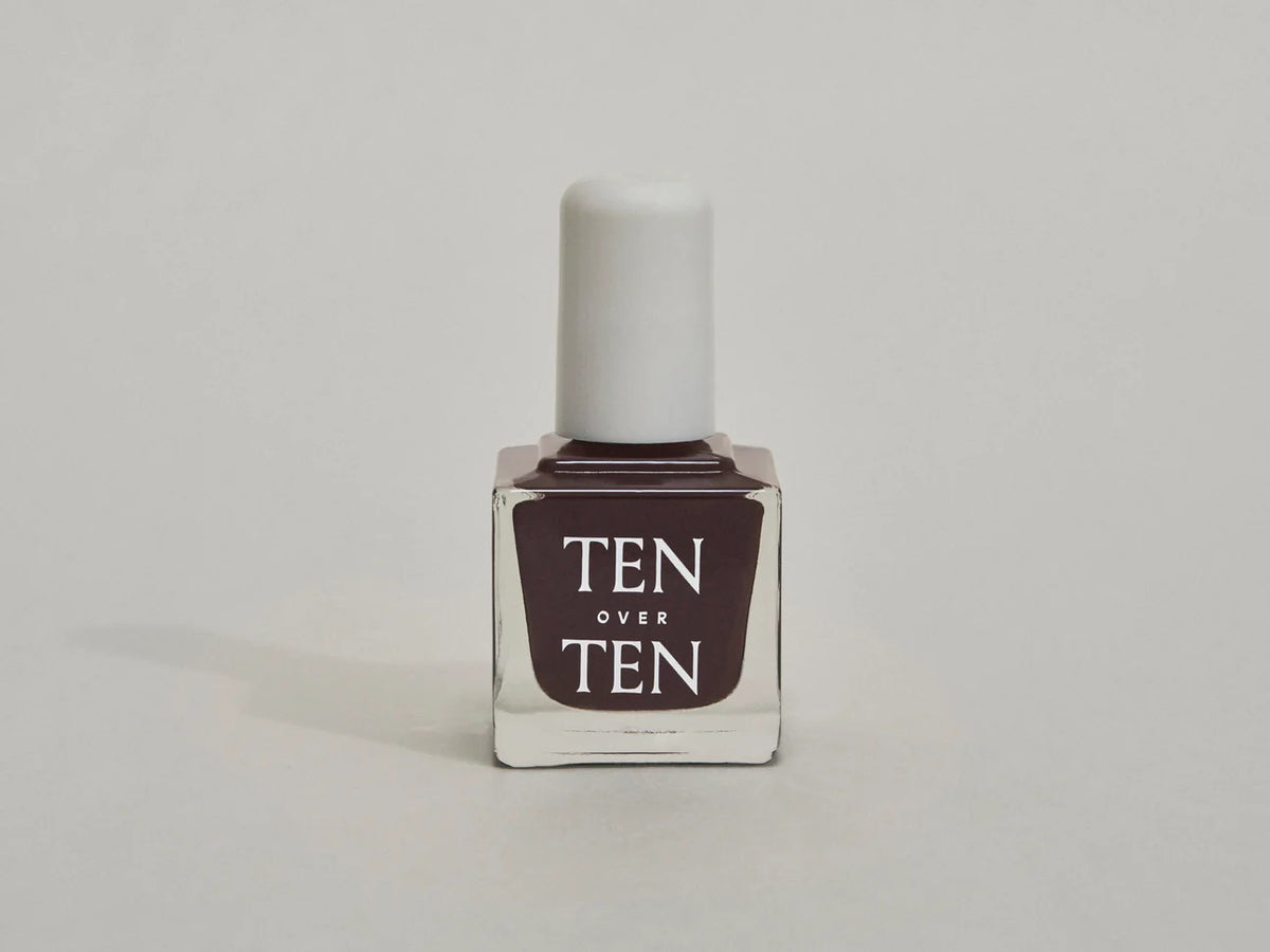 A glass bottle of TENOVERTEN - NAIL POLISH from the brand TEN OVER TEN, featuring a white cap and a dusty pink nail color. The front label displays "TEN OVER TEN" in white uppercase letters. This vegan, eight-free formula ensures both beauty and peace of mind. The background is plain and light-colored.
