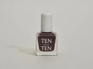 A glass bottle of TENOVERTEN - NAIL POLISH from the brand TEN OVER TEN, featuring a white cap and a dusty pink nail color. The front label displays "TEN OVER TEN" in white uppercase letters. This vegan, eight-free formula ensures both beauty and peace of mind. The background is plain and light-colored.
