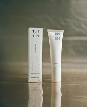 A beige tube labeled "TENOVERTEN - THE HEROINE HAND LOTION" by TEN OVER TEN rests on a clear rectangular platform atop a textured, cracked concrete surface.