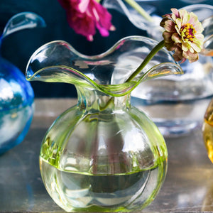 A clear, glass vase with a wide, ruffled opening and a slightly green-tinted round body. Created by Tuscan artisans, the VIETRI Hibiscus Bud Vase Green has a smooth, translucent surface and elegant curves, giving it a modern and artistic appearance.