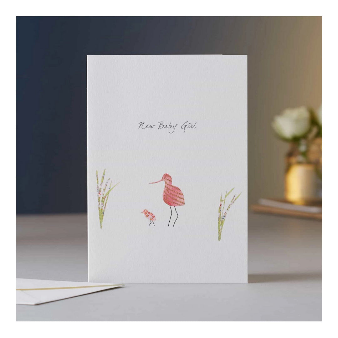 The ELOISE HALL - AVOCET BABY GIRL CARD from ELOISE HALL LTD features the text "New Baby Girl" and a minimalist illustration of two birds, one larger and one smaller, among sprigs of grass. This luxury stationery piece is displayed on a surface with a blurry background that includes a white flower in a vase.
