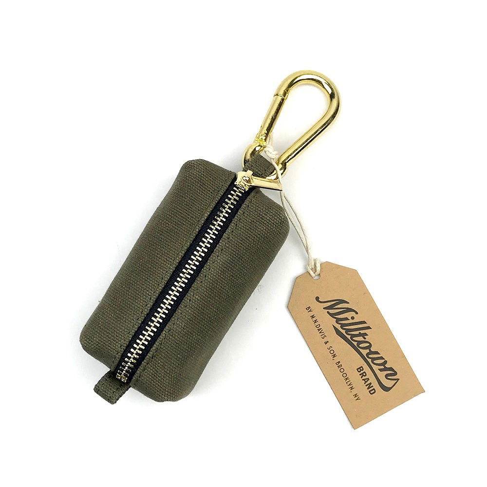 The DOG WASTE DISPENSER (OLIVE) by MILLTOWN BRAND is a small, rectangular pouch made from 12 oz waxed canvas. It features a gold-colored metal zipper and a matching gold carabiner clip, perfect for holding dog waste bags. The pouch includes a tag labeled &quot;MILLTOWN BRAND&quot; with additional information in smaller text.
