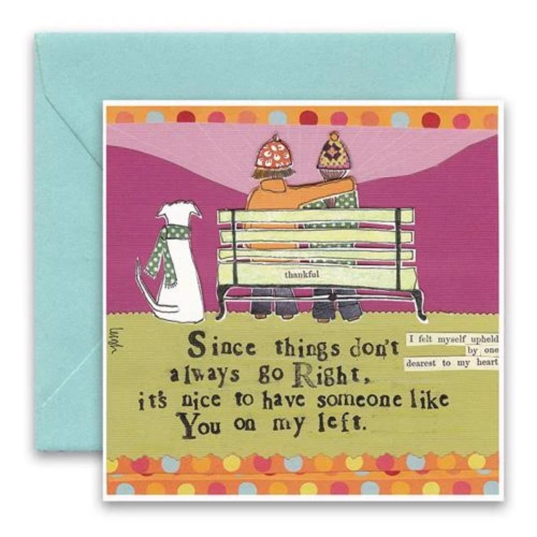 The CURLY GIRL DESIGNS - ON MY LEFT CARD by CURLY GIRL DESIGN, INC features a charming collage art illustration of two people sitting on a bench with a dog to their left. Above them is the heartfelt quote: "Since things don't always go Right, it’s nice to have someone like You on my left." The card is beautifully paired with a teal envelope.