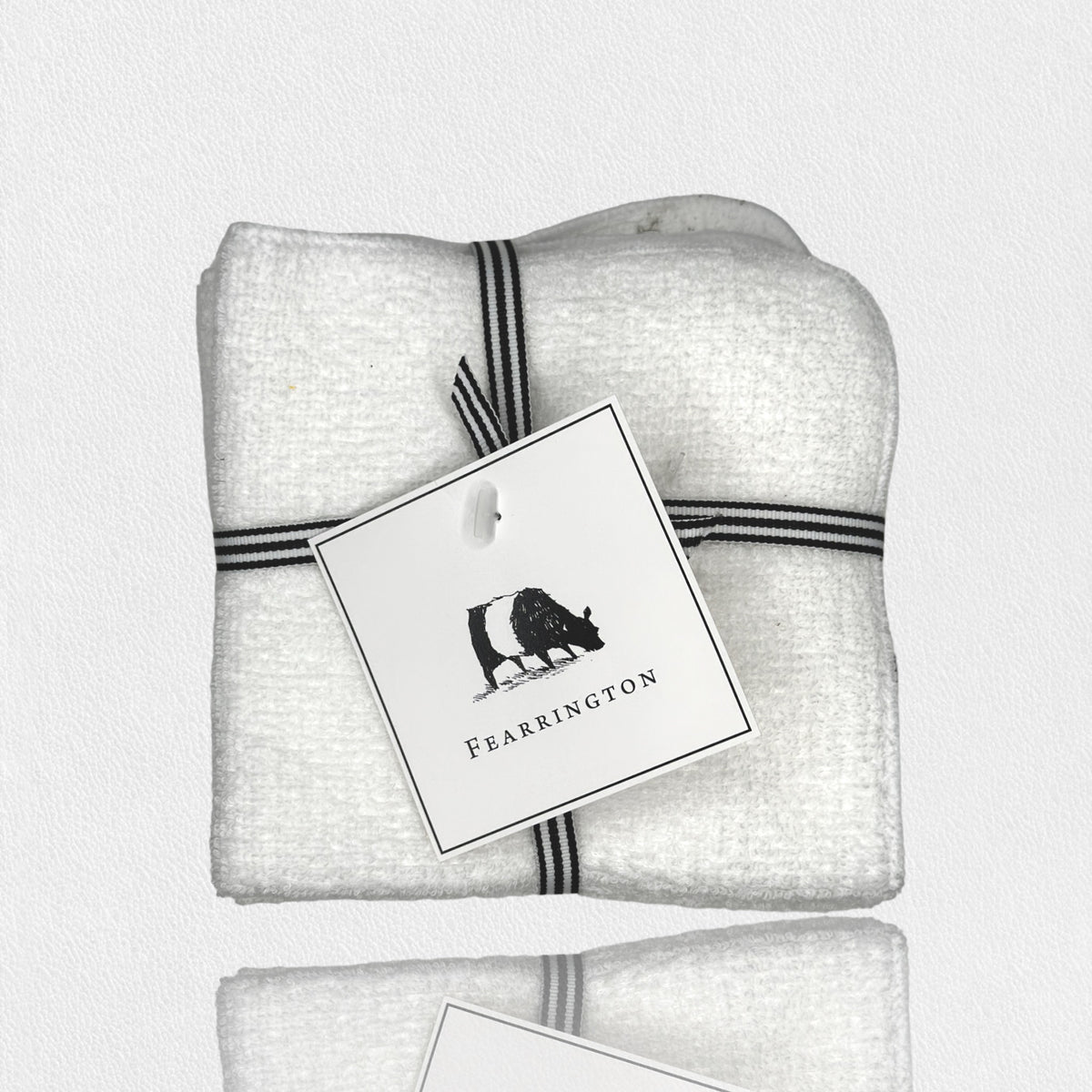 A neatly folded white textured towel, part of the FEARRINGTON LIFESTYLE TOWEL COLLECTION - WAFFLE FACE CLOTH S/4 by HARBOR LINEN, is tied with a black and white striped ribbon. Attached to the ribbon is a square tag featuring a black silhouette of a cow with &quot;FEARRINGTON&quot; in capital letters beneath it. The towel sits on a white surface.