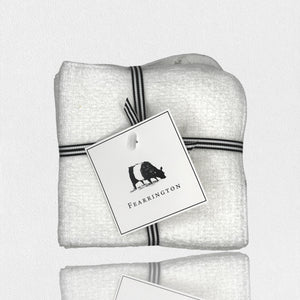 A neatly folded white textured towel, part of the FEARRINGTON LIFESTYLE TOWEL COLLECTION - WAFFLE FACE CLOTH S/4 by HARBOR LINEN, is tied with a black and white striped ribbon. Attached to the ribbon is a square tag featuring a black silhouette of a cow with "FEARRINGTON" in capital letters beneath it. The towel sits on a white surface.