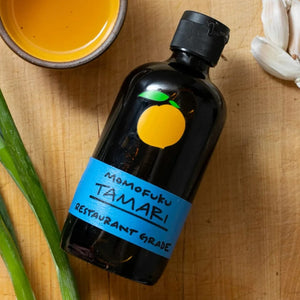 A brown glass bottle with a black cap contains MOMOFUKU - TAMARI, a restaurant-grade, gluten-free soy sauce made from organic ingredients. The label is blue with a minimalist orange fruit logo. The bottle is reflected on the white surface it sits on.