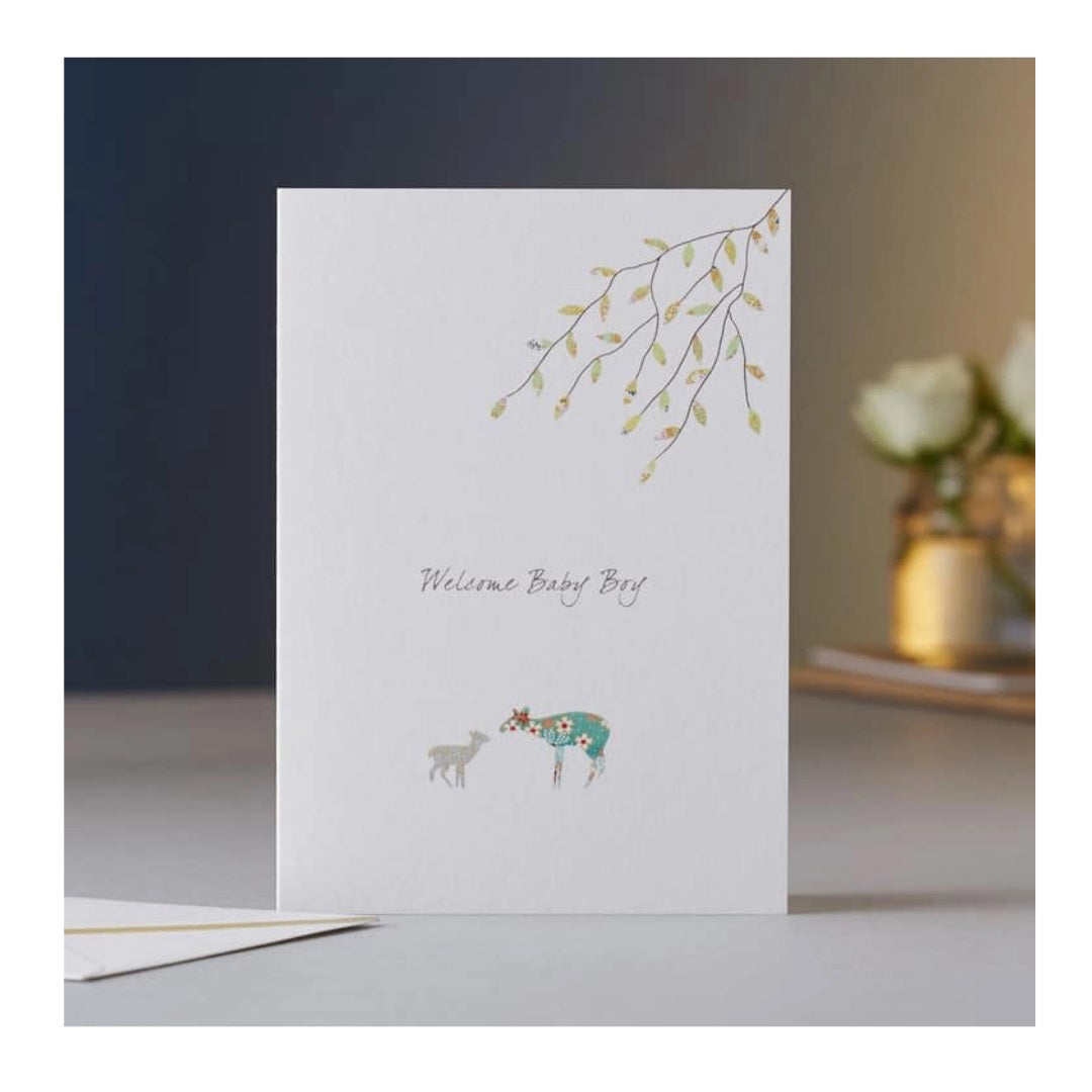 The ELOISE HALL - NEW BABY BOY CARD by Eloise Hall Ltd showcases "Welcome Baby Boy" text prominently in the center. It features a charming illustration of a mother deer and her baby nuzzling, along with an elegant branch and leaves design in the top right corner. The softly blurred background captures the essence of luxury stationery.