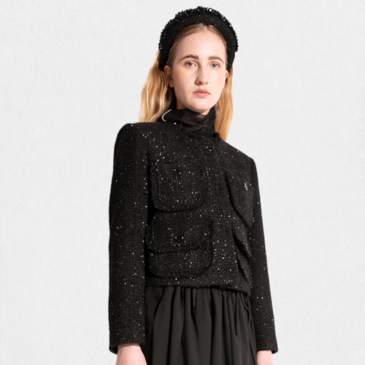 A woman with long blonde hair wears a MEIMEIJ LUREX TWEED JACKET by MEIMEIJ, which features front closure buttons and two large pockets on the chest. She has a black headband and a long black skirt. The background is plain off-white.