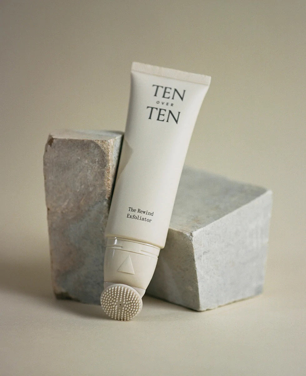 A tube of TENOVERTEN - THE REWIND HAND EXFOLIATOR by TEN OVER TEN, a gentle hand exfoliant with a textured applicator tip, is propped against two gray stone blocks on a neutral background.