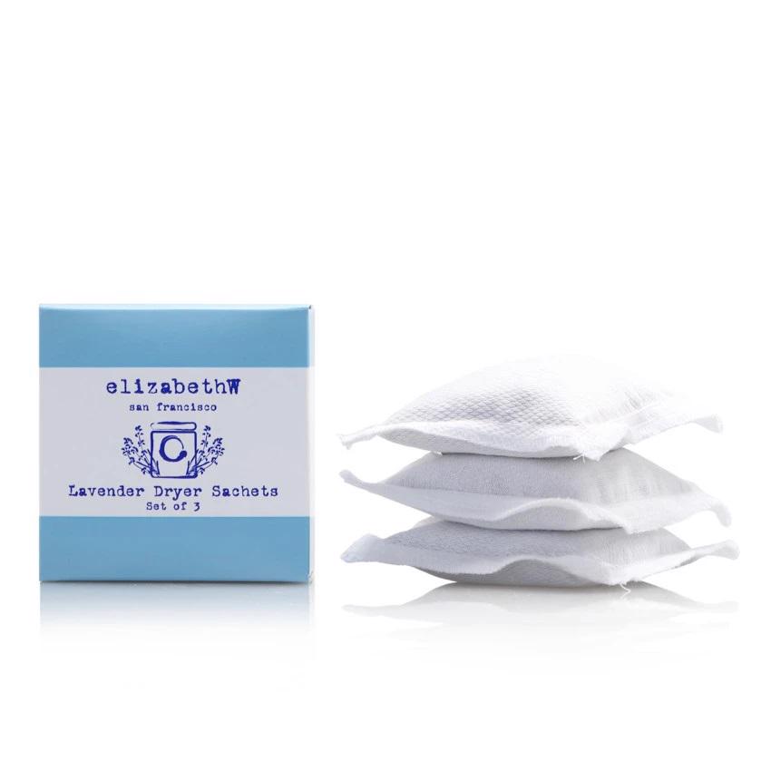 The image depicts a trio of white cloth sachets stacked beside a blue and white box labeled &quot;E WIGHTMAN &amp; CO, LAVENDER DRYER SACHETS, Set of 3.&quot; These eco-friendly pouches appear to contain lavender, likely intended for use in a clothes dryer to impart a gentle lavender fragrance.