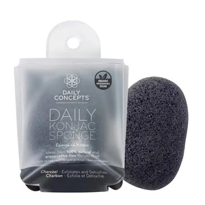 A black Daily Concepts Charcoal Konjac Sponge is placed beside its packaging, labeled "Daily Concepts Daily Konjac Sponge." The packaging highlights that it is made from 100% natural and preservative-free konjac root and features the word "charcoal." It is organic, vegan, biodegradable, and highly effective for removing skin impurities.