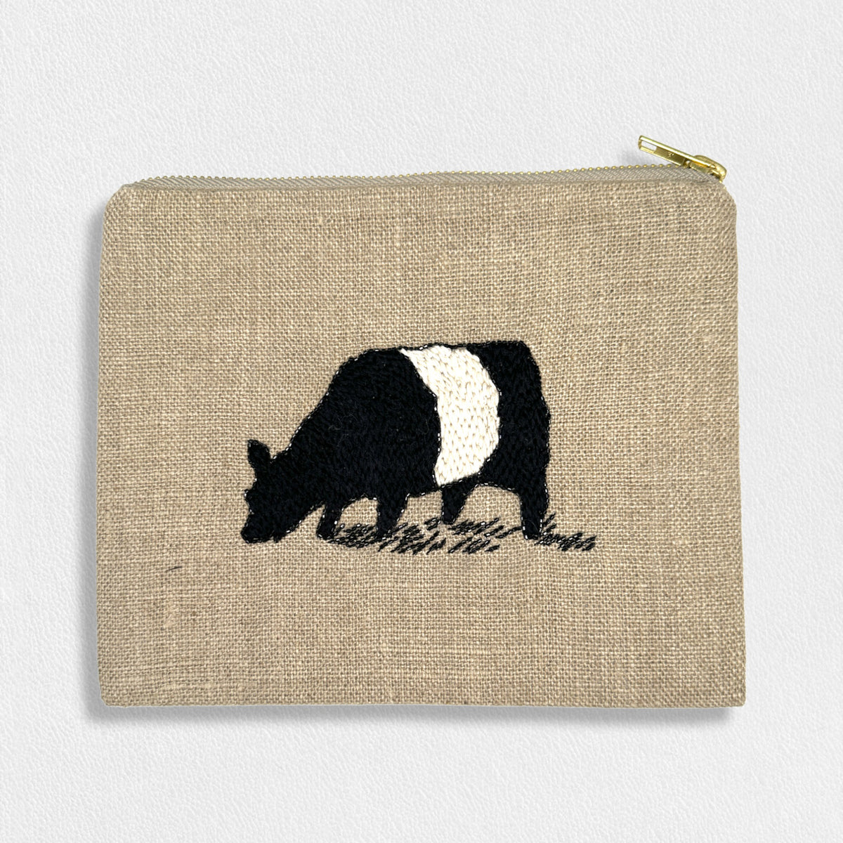 Introducing the Fearrington Lifestyle Collection&#39;s Signature Beltie Linen Pouch by Citrus Fashions: a rectangular beige pouch with a gold zipper. This beautifully crafted pouch showcases an embroidered black and white cow grazing on grass in the center, highlighting hand-stitched details. The natural, textured fabric is perfect for holding light cosmetics.