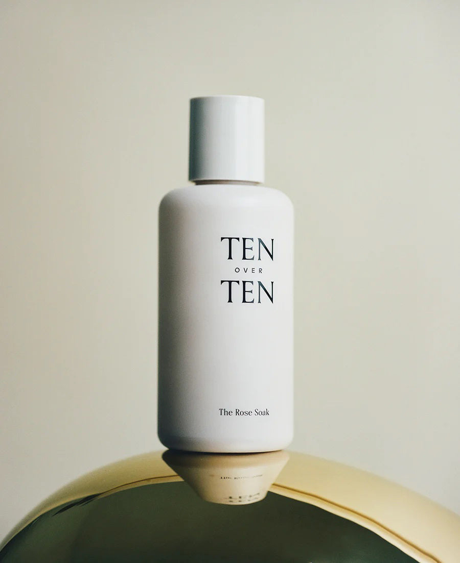 A white bottle labeled "TENOVERTEN - THE ROSE SOAK POLISH REMOVER" by TEN OVER TEN stands upright on a reflective gold surface, set against a neutral background.