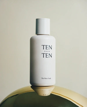 A white bottle labeled "TENOVERTEN - THE ROSE SOAK POLISH REMOVER" by TEN OVER TEN stands upright on a reflective gold surface, set against a neutral background.