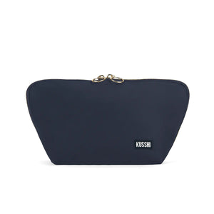 The KUSSHI Signature Makeup Bag in Navy features a minimalist design with a slightly triangular shape. This navy blue zipped pouch includes a black rectangular label with white text that reads "KUSSHI" near the bottom right corner and two golden pull tabs at the top center. Additionally, this makeup bag is machine washable.