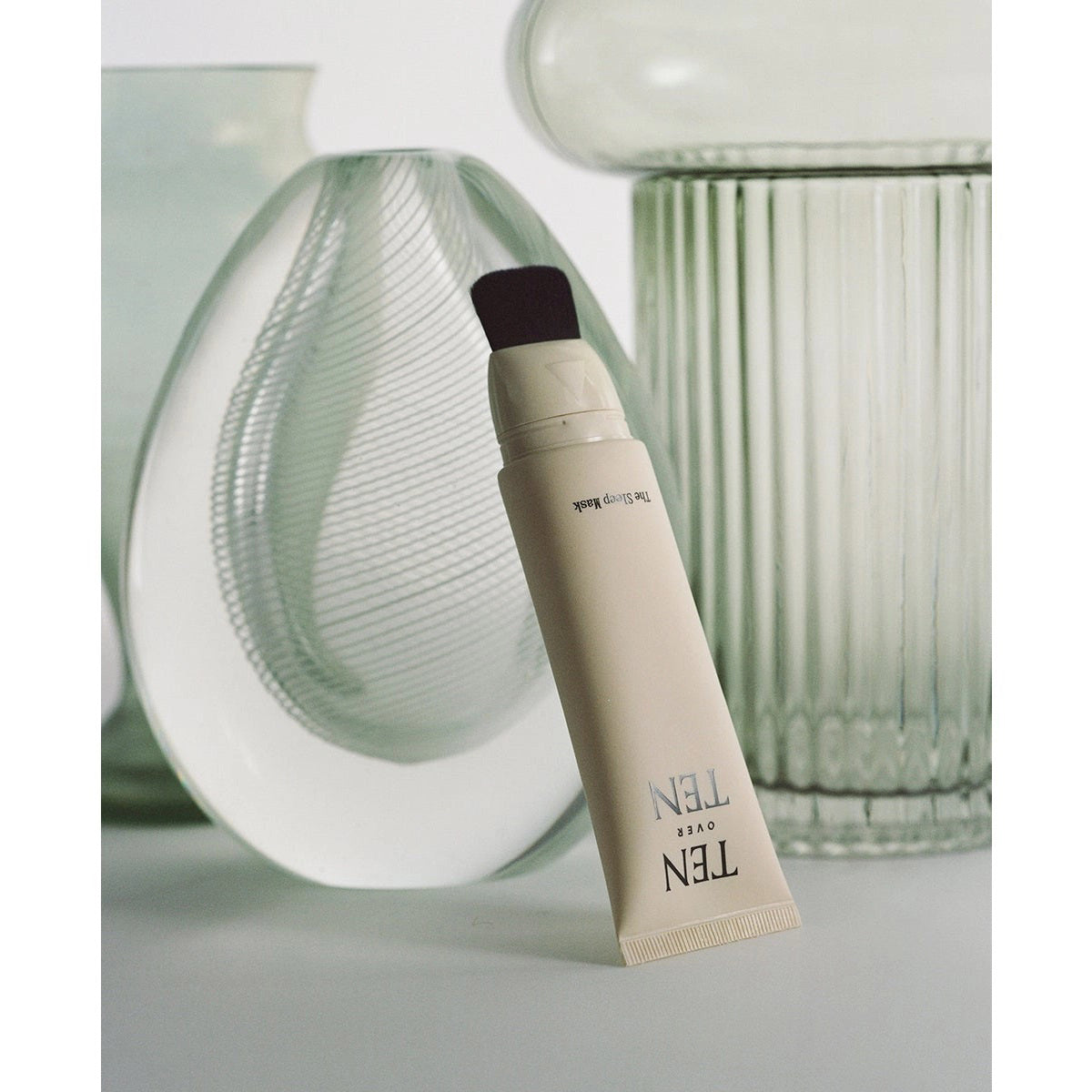 A beige tube labeled "TEN OVER TEN - THE SLEEP MASK" is displayed horizontally in front of textured glass vases on a soft, light background—an elegant way to showcase this luxurious hand cream from TEN OVER TEN.