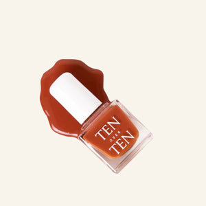 A glass bottle of TENOVERTEN - NAIL POLISH from the brand TEN OVER TEN, featuring a white cap and a dusty pink nail color. The front label displays "TEN OVER TEN" in white uppercase letters. This vegan, eight-free formula ensures both beauty and peace of mind. The background is plain and light-colored.
