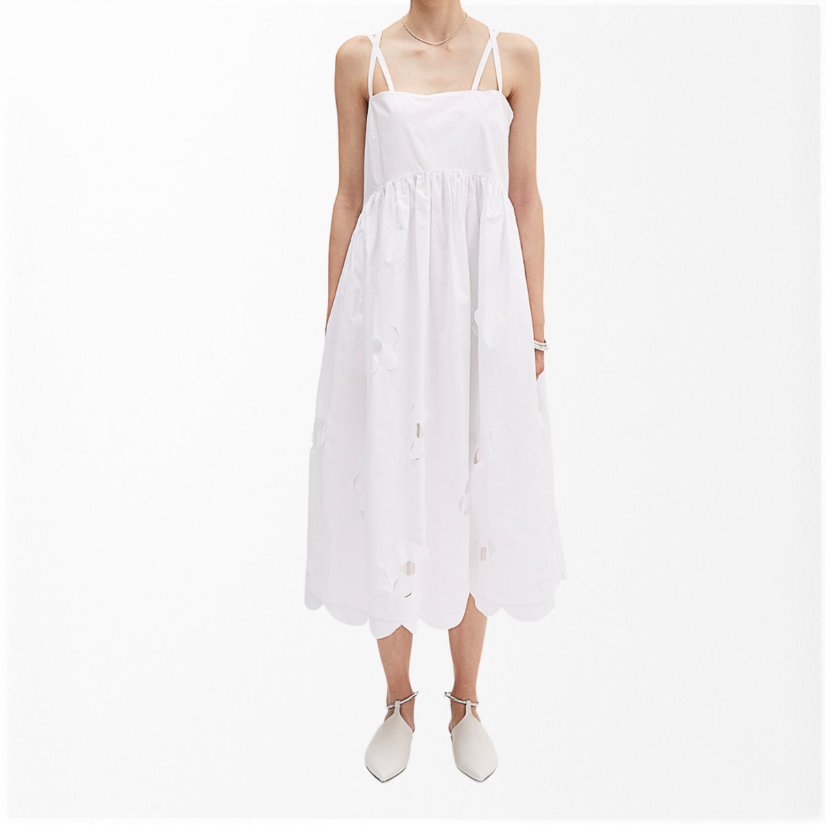 A person wearing the MEIMEIJ Cotton Midi Dress, featuring sleeveless design with delicate cut-out floral patterns, stands against a plain white background. The light dress from MEIMEIJ includes criss-cross straps, a gathered waist, and a scalloped hemline. They are also wearing white shoes with pointed toes.