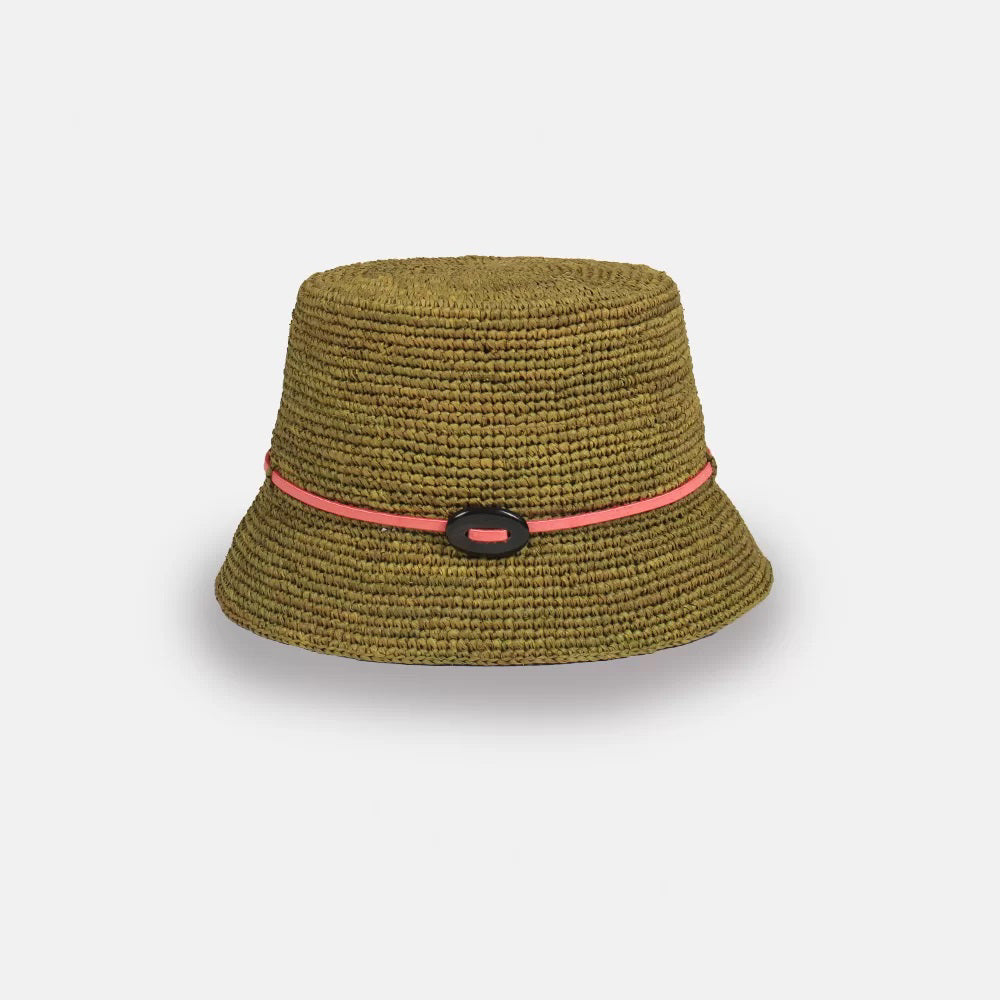 The LEO BUCKET HAT by SANS ARCIDET is a handmade crocheted bucket hat in pink featuring a slightly flared brim. This women's accessory boasts a thin blue cord woven around the base and an oval-shaped black decoration at the front. Crafted from natural raffia, it stands out beautifully against the plain white background.