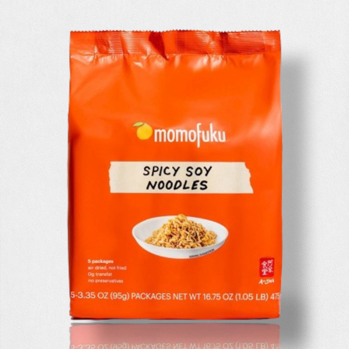 A vibrant orange package of MOMOFUKU Spicy Soy Noodles highlights savory and spicy flavors. The front features the MOMOFUKU brand logo, a small white bowl of noodles, and various product details. It includes five individual packs of air-dried noodles, each weighing 95 grams, with a total weight of 475 grams.