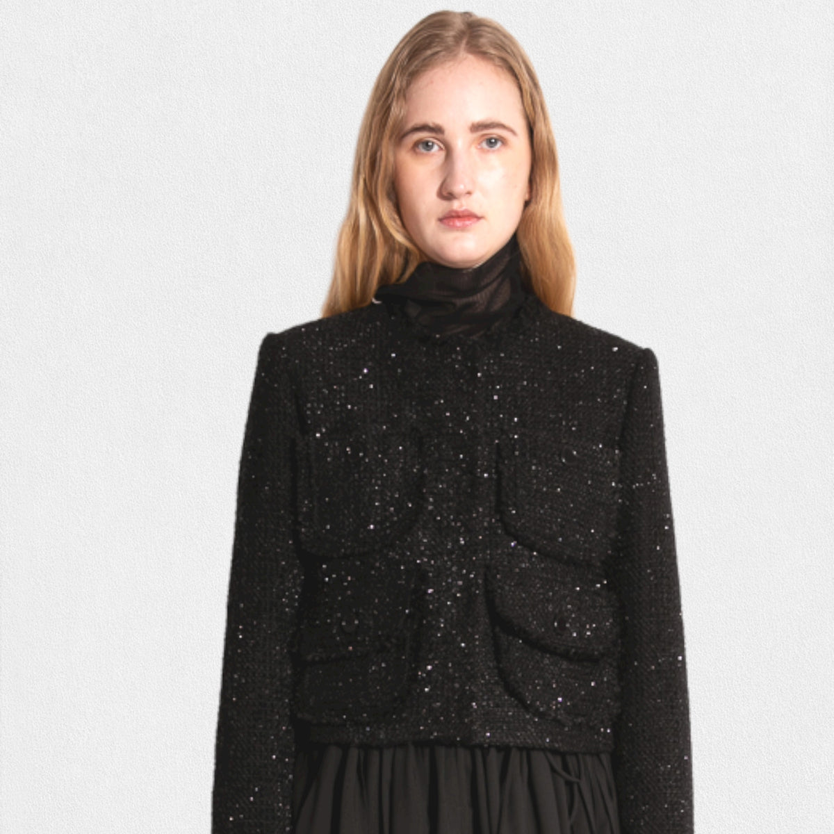 A woman with long blonde hair wears a MEIMEIJ LUREX TWEED JACKET by MEIMEIJ, which features front closure buttons and two large pockets on the chest. She has a black headband and a long black skirt. The background is plain off-white.