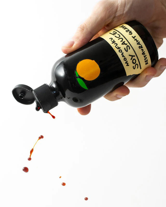 A dark brown bottle with a black cap, featuring a label with an orange fruit graphic at the top. Below the graphic, the label reads "MOMOFUKU - SOY SAUCE" and "Restaurant Grade" in bold, handwritten-style text—perfect for home cooks who value organic ingredients from MOMOFUKU.