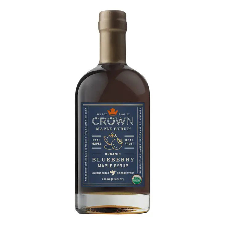 A glass bottle of CROWN MAPLE - BLUEBERRY MAPLE SYRUP from Crown Maple, featuring a gold cap and a navy blue label adorned with gold and white text along with blueberry imagery, contains 250 mL (8.5 fl oz) of Pure Organic Maple Syrup. The label emphasizes "Real Maple" and "Real Fruit.