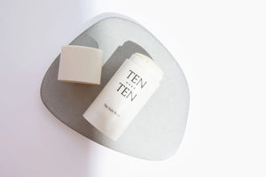 An open white cylindrical container displaying the brand "TEN OVER TEN" and product name "TENOVERTEN - THE SOLE MATE." The lid rests beside the container, revealing a solid cream stick enriched with botanical oils. The minimalist, light-colored background accentuates how effectively you can moisturize feet.