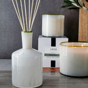 A white ceramic diffuser bottle with reed sticks is displayed in front of a minimalistic, white rectangular box. The box features a vertically centered black ribbon and a label that reads, "LAFCO New York, Feu de Bois, Sandalwood, Leather, Virginia Cedarwood." This LAFCO Ski House Feu de Bois Diffuser is perfect for creating a warm ambiance.