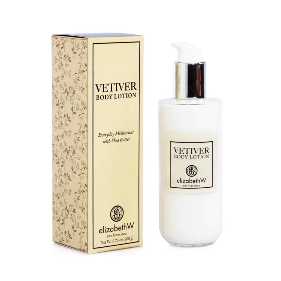 A bottle of E Wightman &amp; Co Vetiver Body Lotion is placed next to its decorative packaging box. The bottle, featuring a pump dispenser and a label indicating it contains an everyday moisturizer with shea butter and vitamin E, promises optimal hydration. The box has a floral pattern and similar labeling.