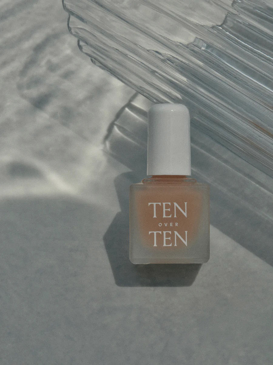 A bottle of TENOVERTEN – THE FOUNDATION BASE COAT by TEN OVER TEN, featuring a white cap, rests on a light textured surface with soft shadows and glass reflections, ideal for supporting healthy natural nails.