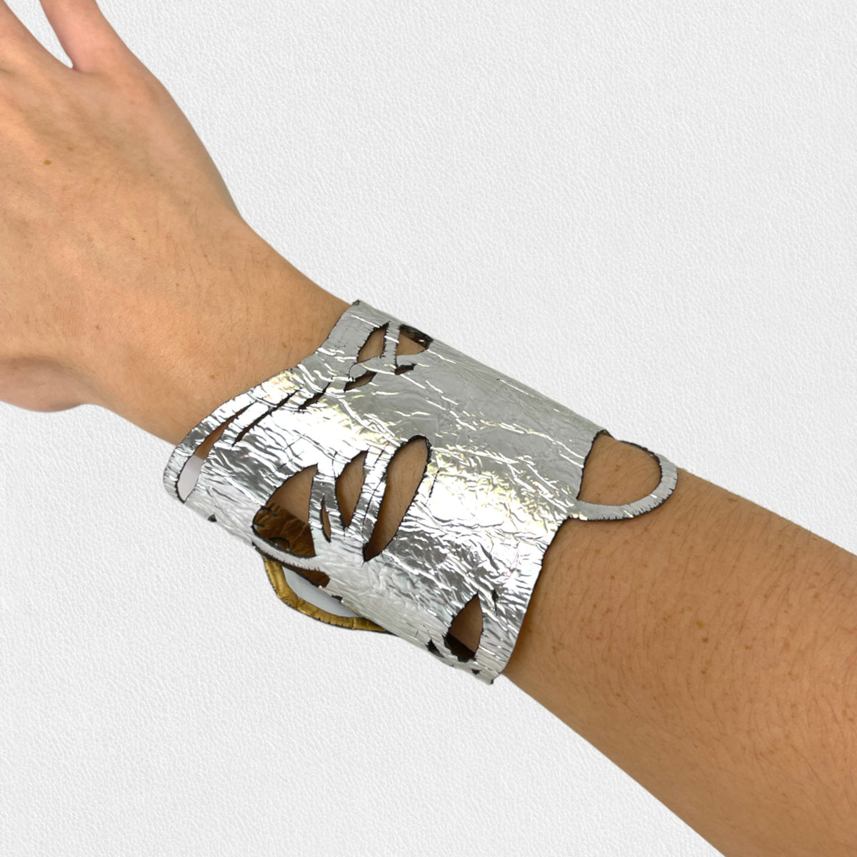 A person wearing the ILLUSIONISM BRACELET IN GOLD AND SILVER (REVERSIBLE) from SI SABATO ISABEL on their wrist. The handmade jewellery, crafted by an Argentinian artist, features irregular cut-out shapes and an intricate, hammered surface. The person is dressed in a faded blue denim shirt with white buttons.