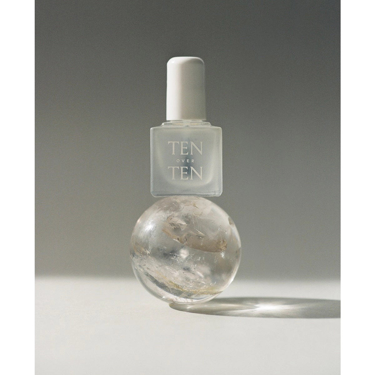 A frosted glass bottle labeled "TEN OVER TEN" with a white cap, containing TENOVERTEN - THE SHIELD TOP COAT, is balanced on a clear spherical object against a plain, softly lit background.