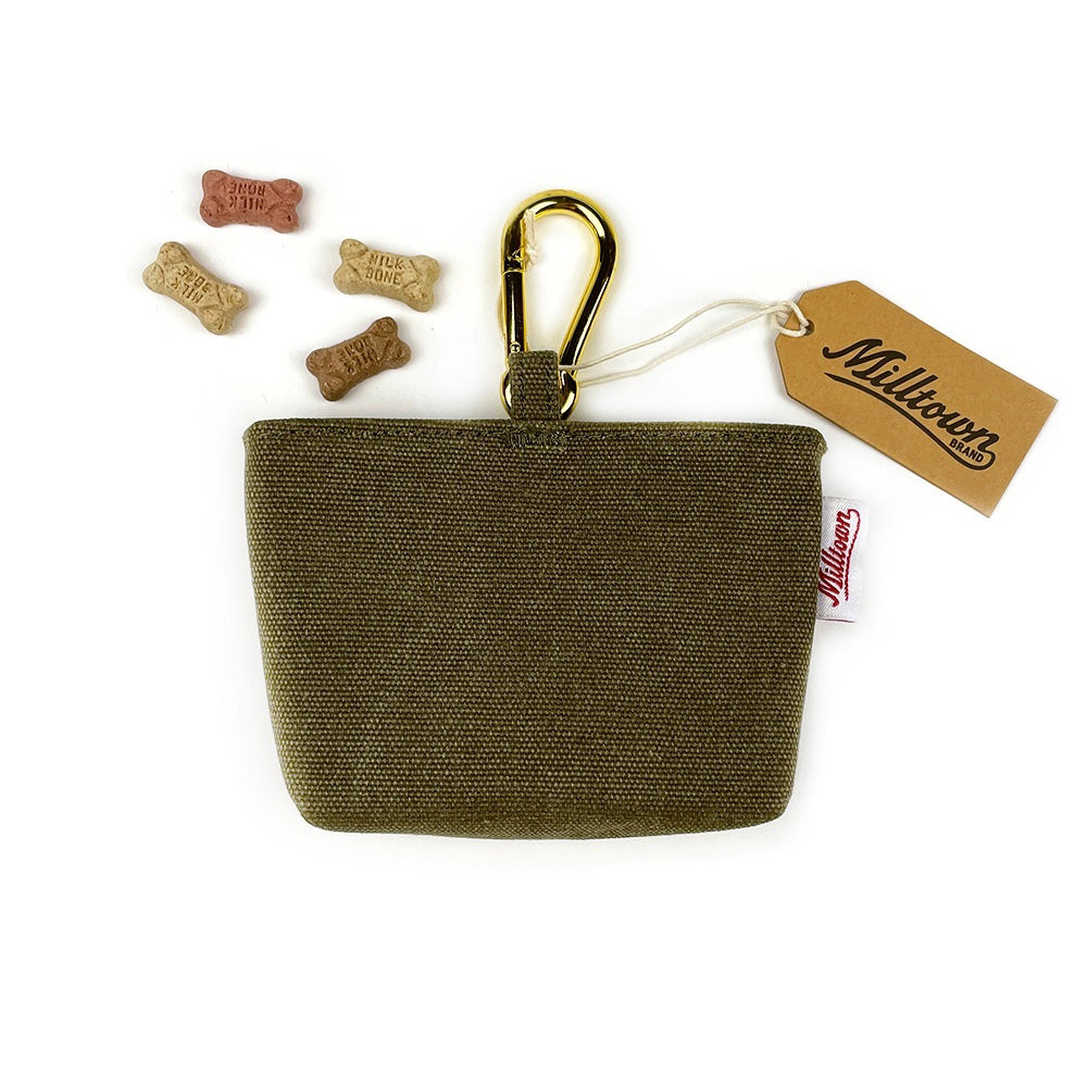 A small, rectangular olive-green DOG TREAT CASE (OLIVE) with a zippered top and a brass clip attached is displayed. A tag with the MILLTOWN BRAND logo is affixed to the clip. Five bone-shaped dog treats in various colors are scattered around the case, which boasts a minimalist design perfect for pet owners on the go.