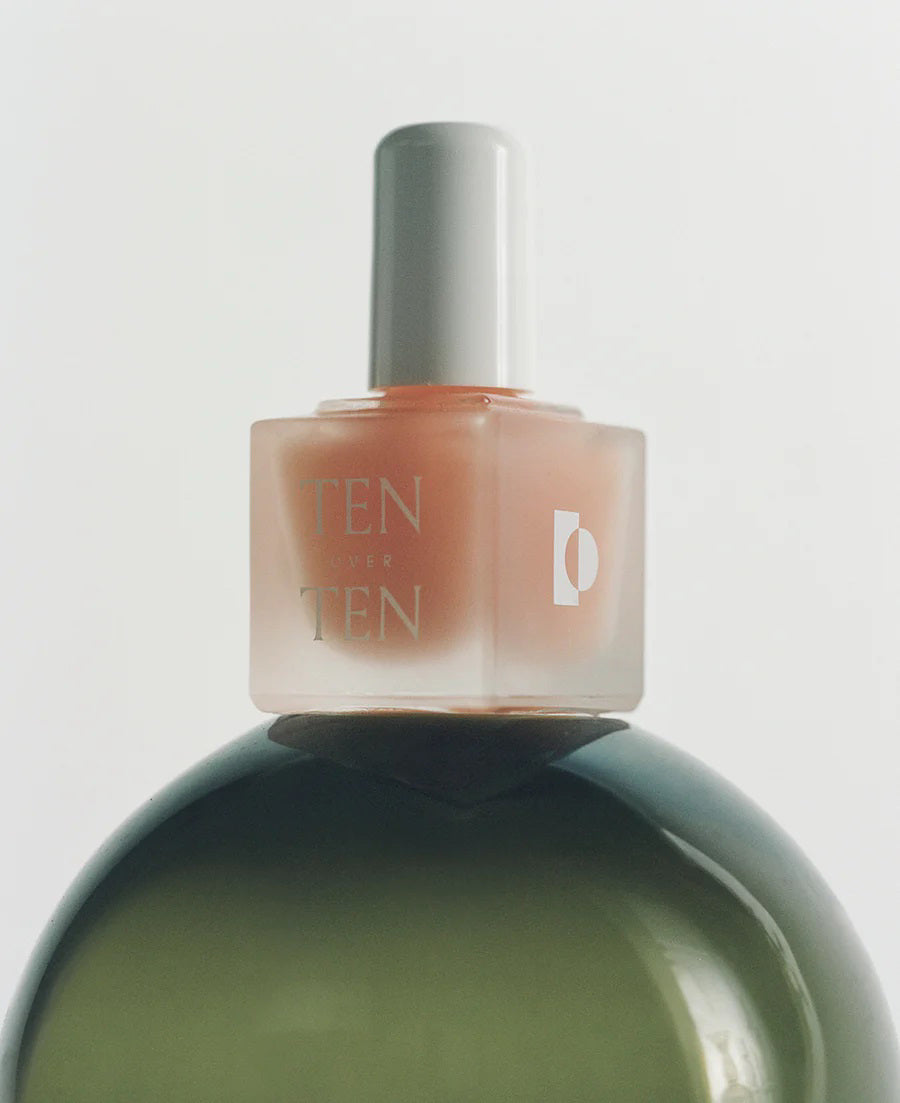 A bottle of TENOVERTEN – THE FOUNDATION BASE COAT by TEN OVER TEN, featuring a white cap, rests on a light textured surface with soft shadows and glass reflections, ideal for supporting healthy natural nails.