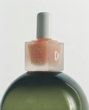 A bottle of TENOVERTEN – THE FOUNDATION BASE COAT by TEN OVER TEN, featuring a white cap, rests on a light textured surface with soft shadows and glass reflections, ideal for supporting healthy natural nails.