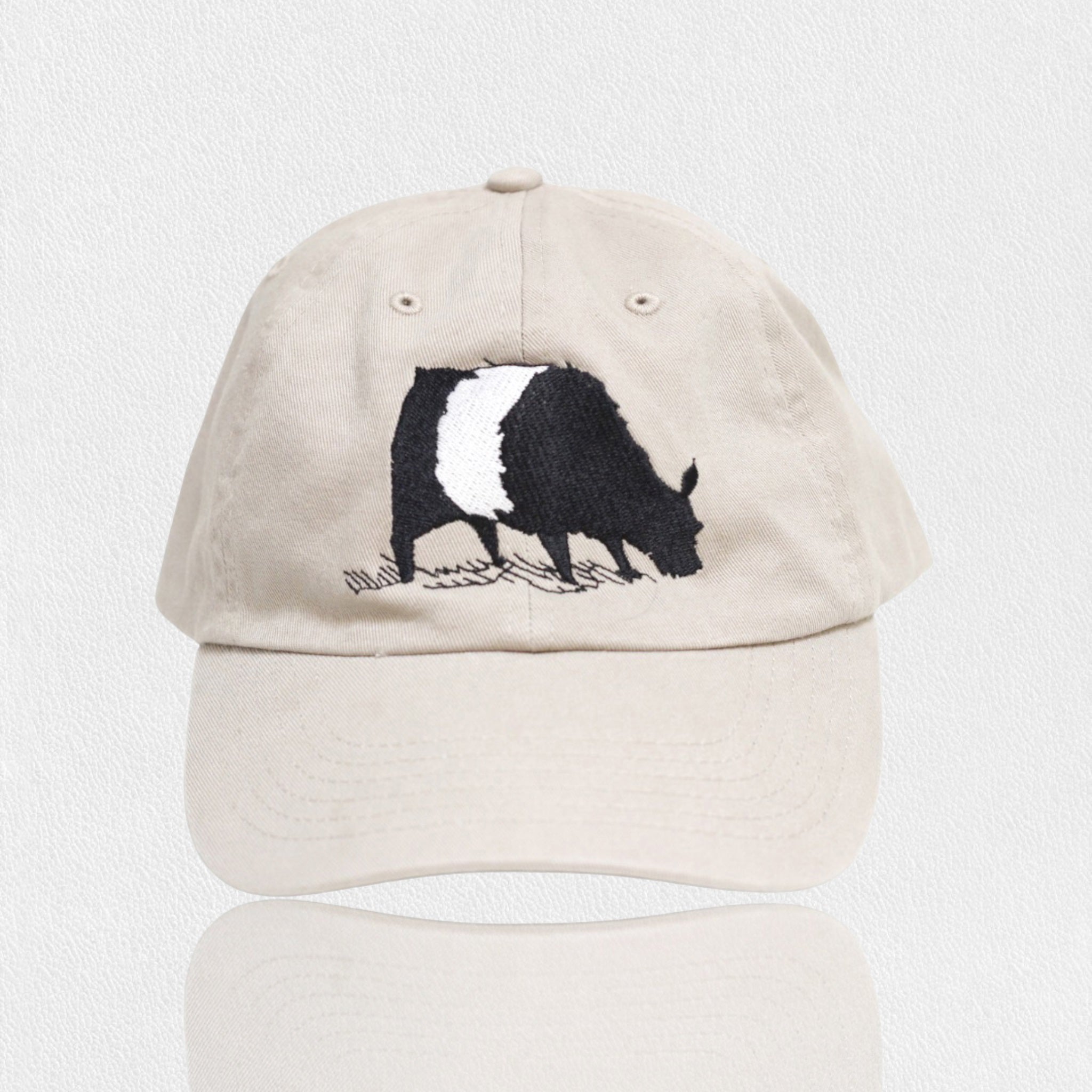 A beige baseball cap featuring an embroidered black and white cow grazing on grass, centered above the brim on the front panel.