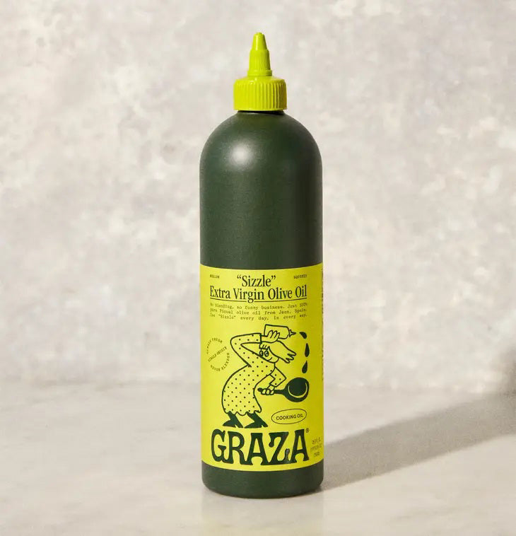 A green bottle with a yellow label containing GRAZA - SIZZLE OLIVE OIL from the brand Graza is placed on a light-colored surface against a neutral background. The label, featuring an illustration of a person pouring olive oil and cooking-related text and graphics, proudly showcases its high-quality picual olive origins.