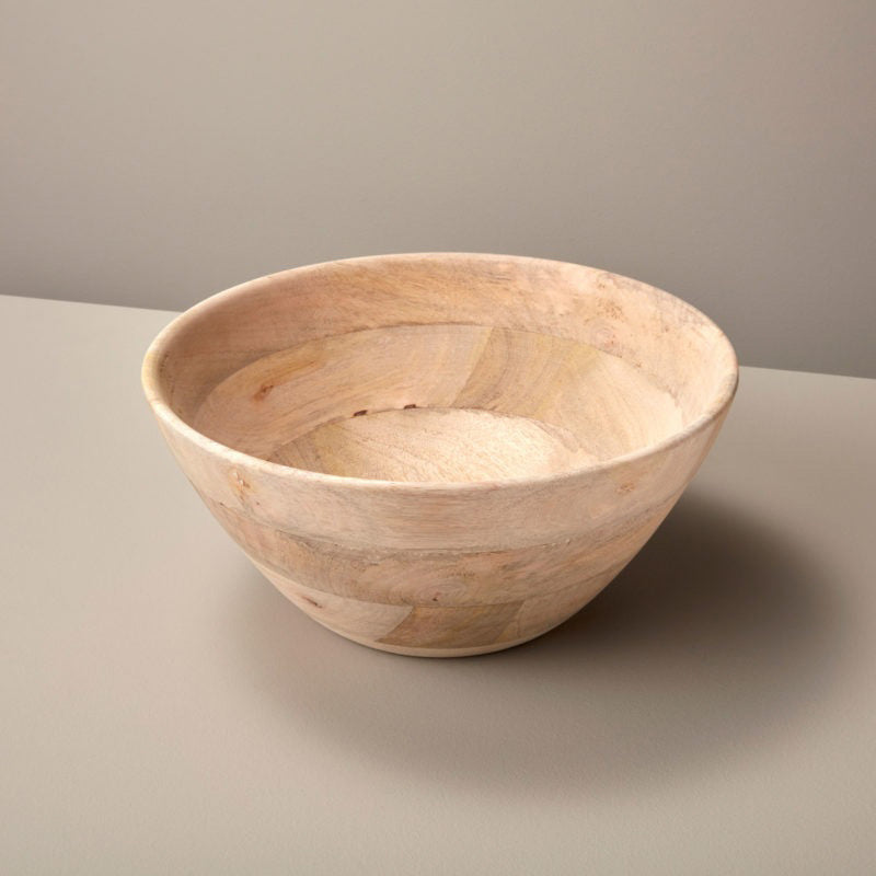 The CORISSA BOWL - LARGE by BE HOME, INC is a hand-carved, round bowl made from sustainably sourced Raw Mango Wood, featuring a smooth matte finish and light wood color, displayed on a neutral gray surface against a plain background.